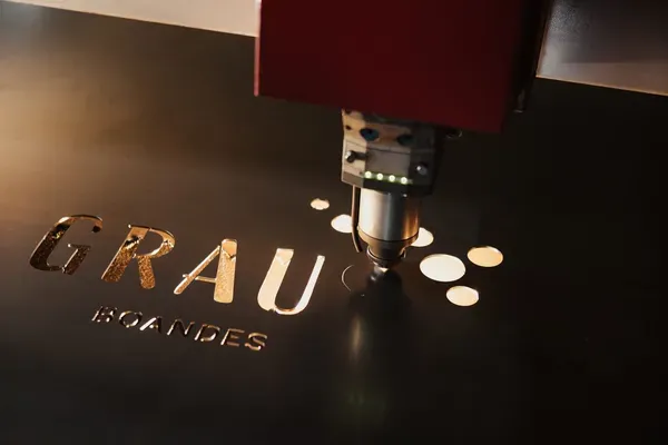 Lasermaschine schneidet Buchstaben 'GRAU' in dunkles Metallblech, neben runden Kreisen; roter Kopf, Südtiroler Metallbaufirma.