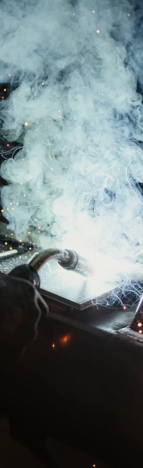 Eine Person schweißt an einer Metallkonstruktion in einem Südtiroler Betrieb; Funken und Rauch umgeben den Lichtbogen.