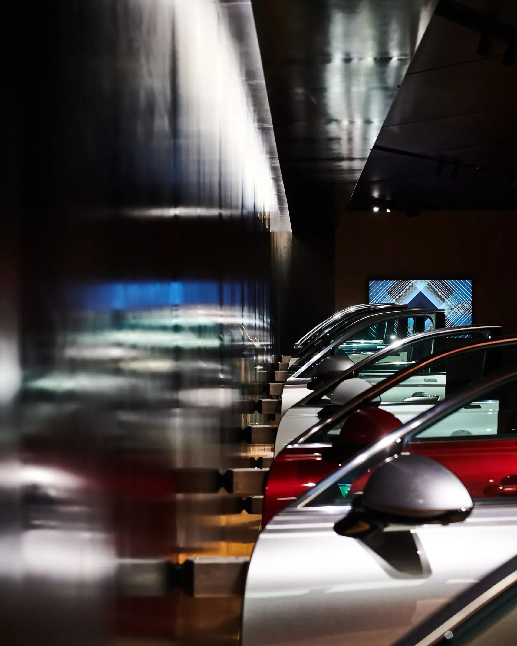 Mehrere Autos in einer dunklen Industrieausstellung, hintereinander geparkt; links reflektierende Metallwand, rechts Fahrzeugfronten sichtbar.